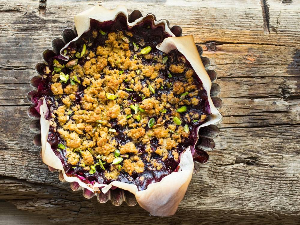 Smuldrepai med bær og pistasj på en treplate, sett fra oven.