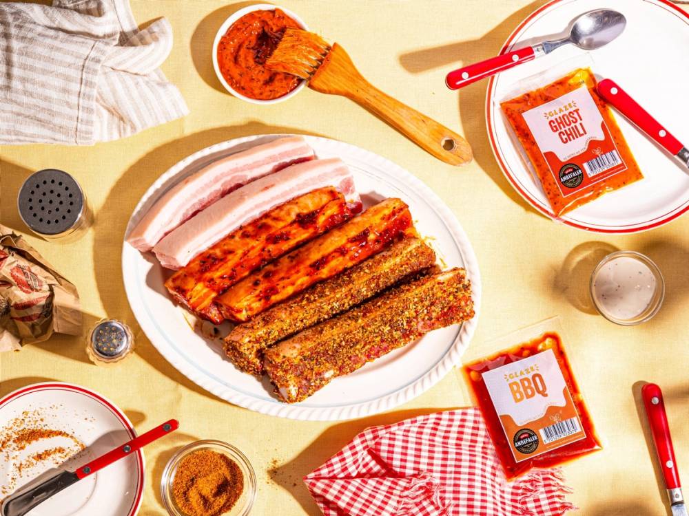 Et fat med ulike typer fersk grillmat, inkludert kjøtt og pølser, omgitt av tilbehør og krydder på et bord med sommerstemning.