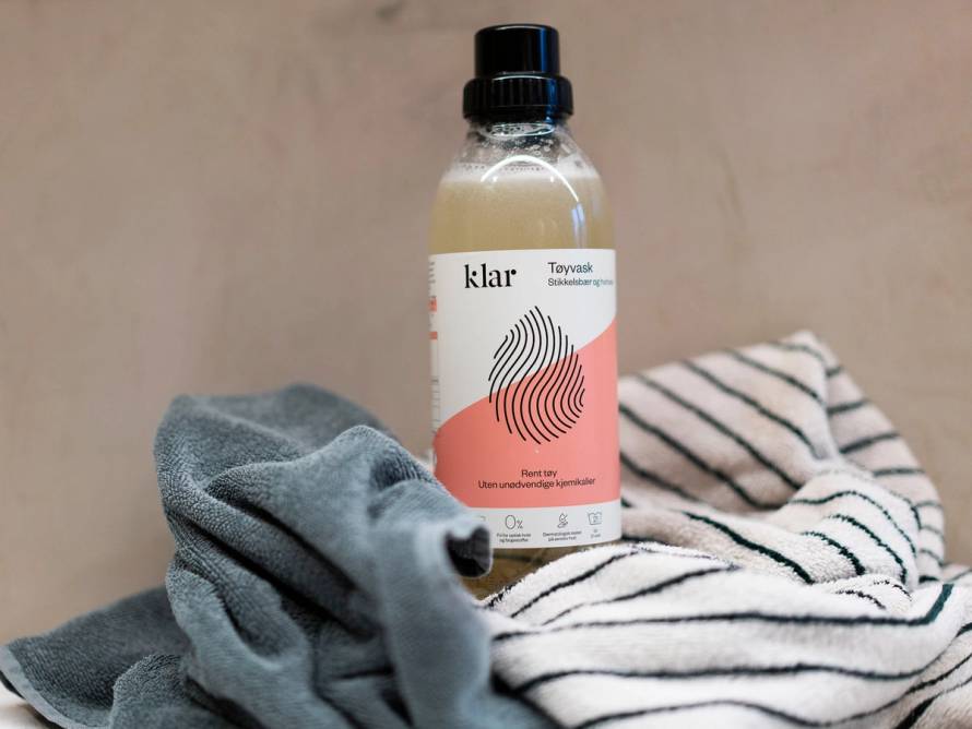 En flaske med vaskemiddel står oppreist på en overflate sammen med klesvask og et håndkle, som antyder at det er snakk om vaskemiddel til tøyvask.
