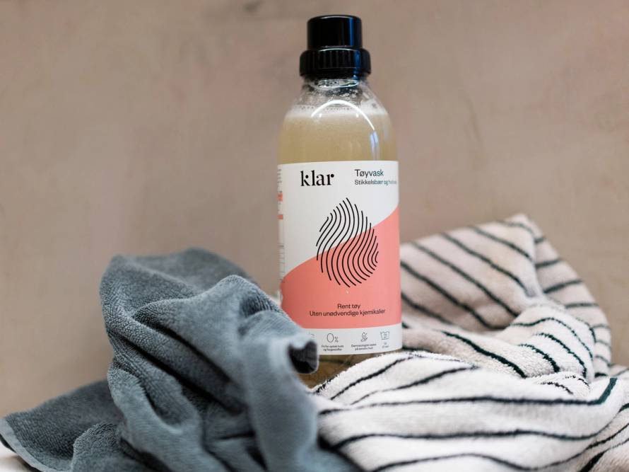 En flaske med vaskemiddel står oppreist på en overflate sammen med klesvask og et håndkle, som antyder at det er snakk om vaskemiddel til tøyvask.
