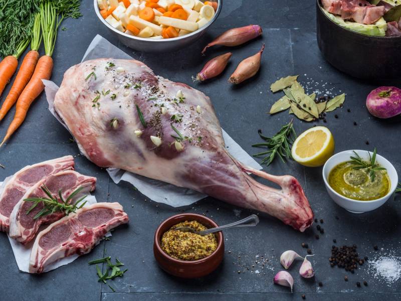 Ulike råvarer til lam, inkludert en hel lammekjøtt, grønnsaker, krydder og sauser, på en mørk stekeplate.