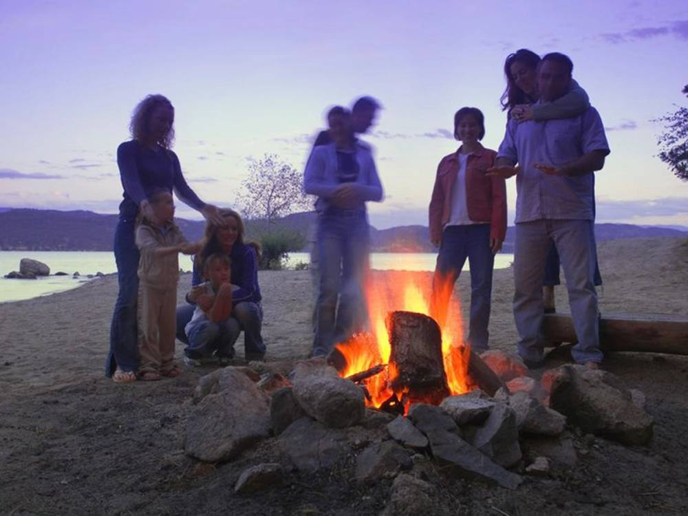 En gruppe mennesker samles rundt et bål ved vannet under solnedgang, med noen som står og snakker og andre som sitter på steiner.