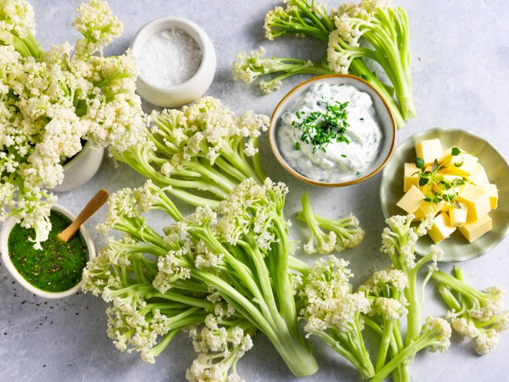 Buketter av blomkål og andre grønnsaker på et kjøkkenbord, med en liten skål med saus og smør i bakgrunnen.