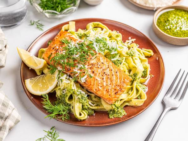 Tagliatelle med pesto og laks servert på en tallerken med sitronbåter og ferske urter, omgitt av små skåler med saus og urter.