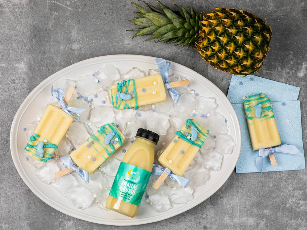 En tallerken med små gaver og en flaske med drikke, omgitt av en ananas, på en grå overflate.