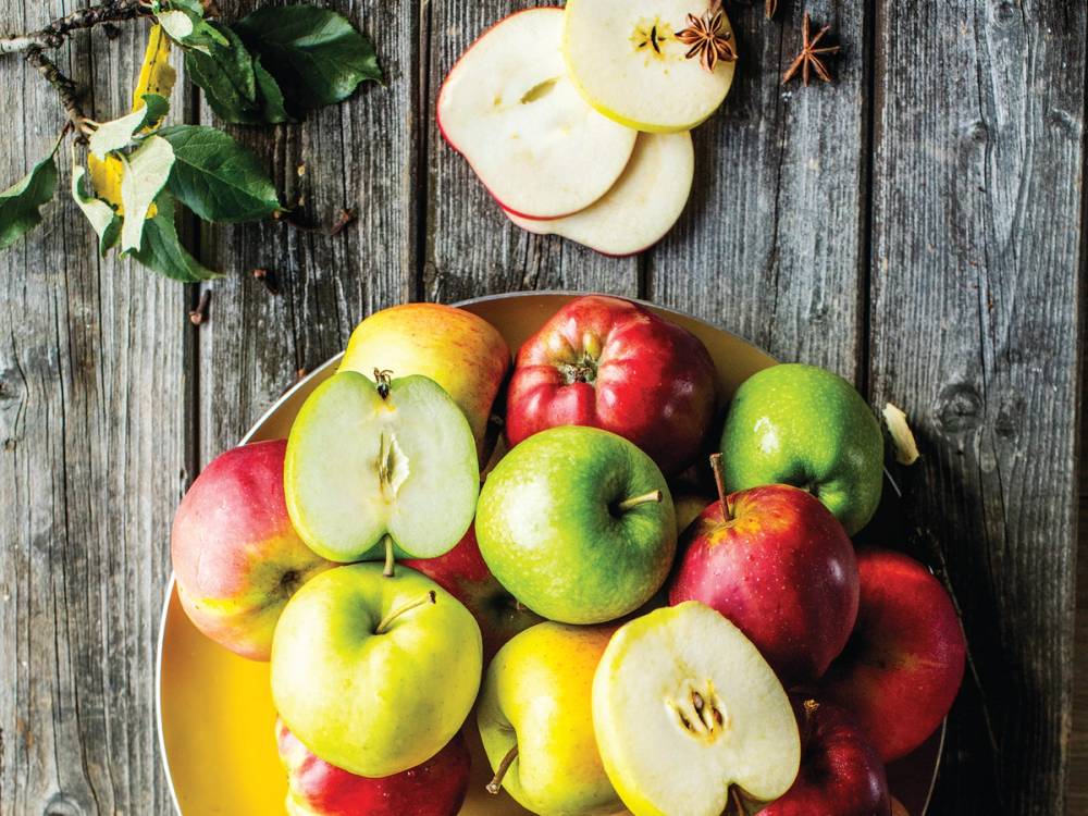 En skål med ulike typer epler, noen hele og noen skåret opp, plassert på et trebord med blader og stjerneanis rundt.