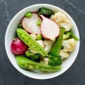 En bol med ulike rå grønnsaker som blomkål, reddik, sukkererter, brokkoli, og grønne erter, på en mørk bakgrunn.