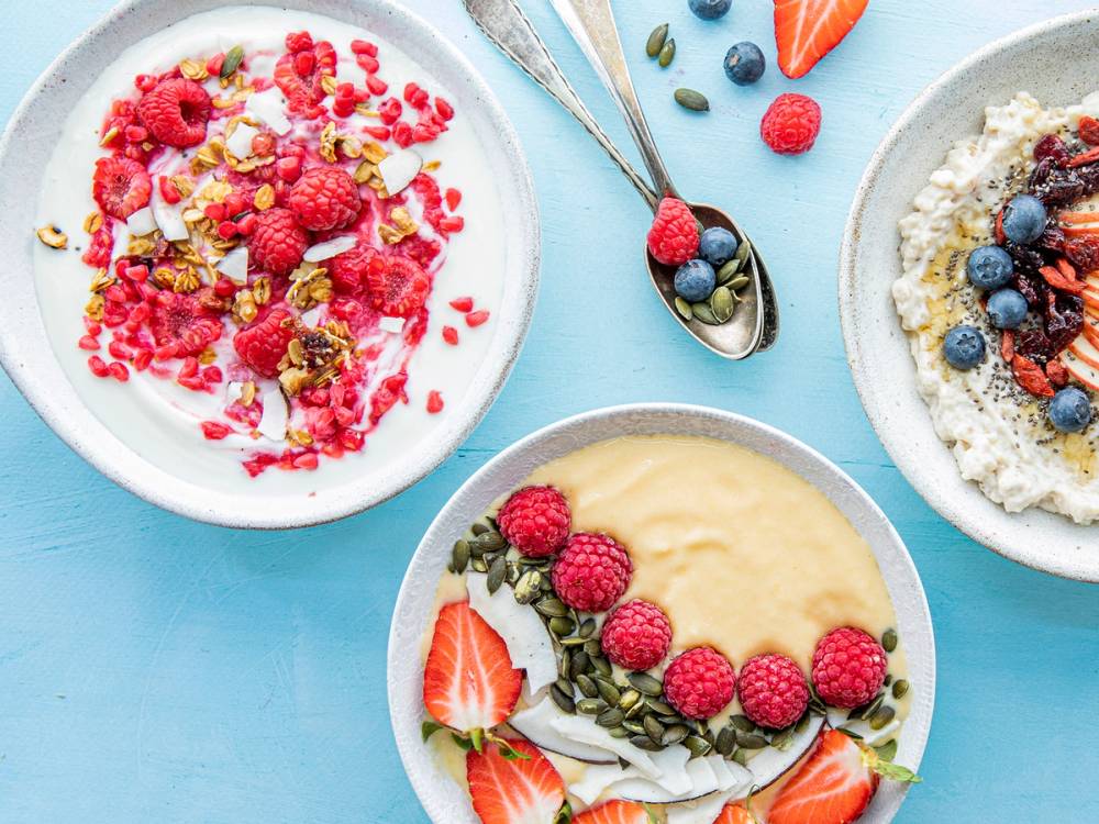 Frokost med bringebær, blåbær og jordbær på ulike tallerkener, med en skje og en skål med yoghurt eller dessert, på en lys bakgrunn.