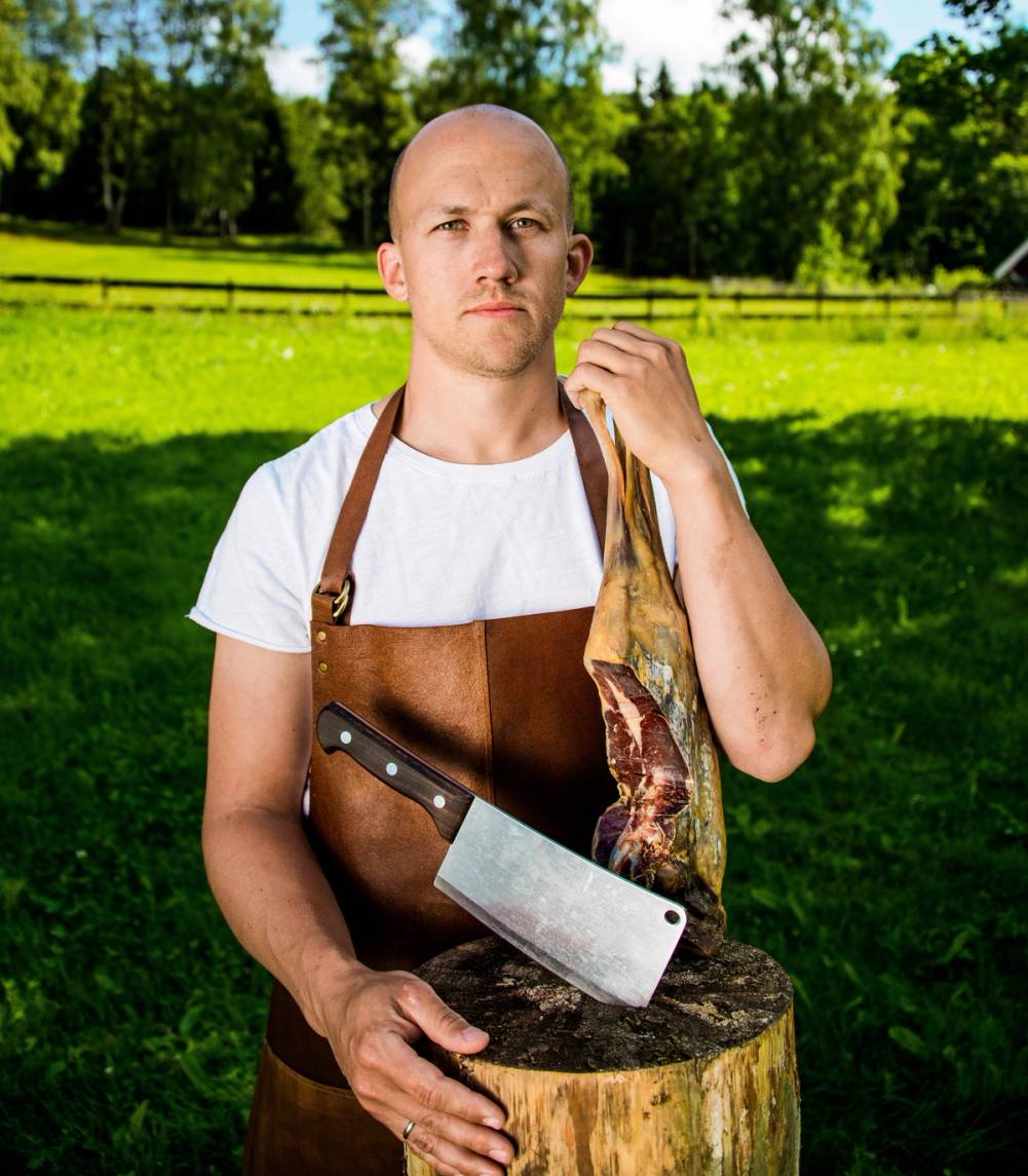 Person med forkle og kniv står ved en trestamme i en grønn hage eller park, med trær og gjerde i bakgrunnen.