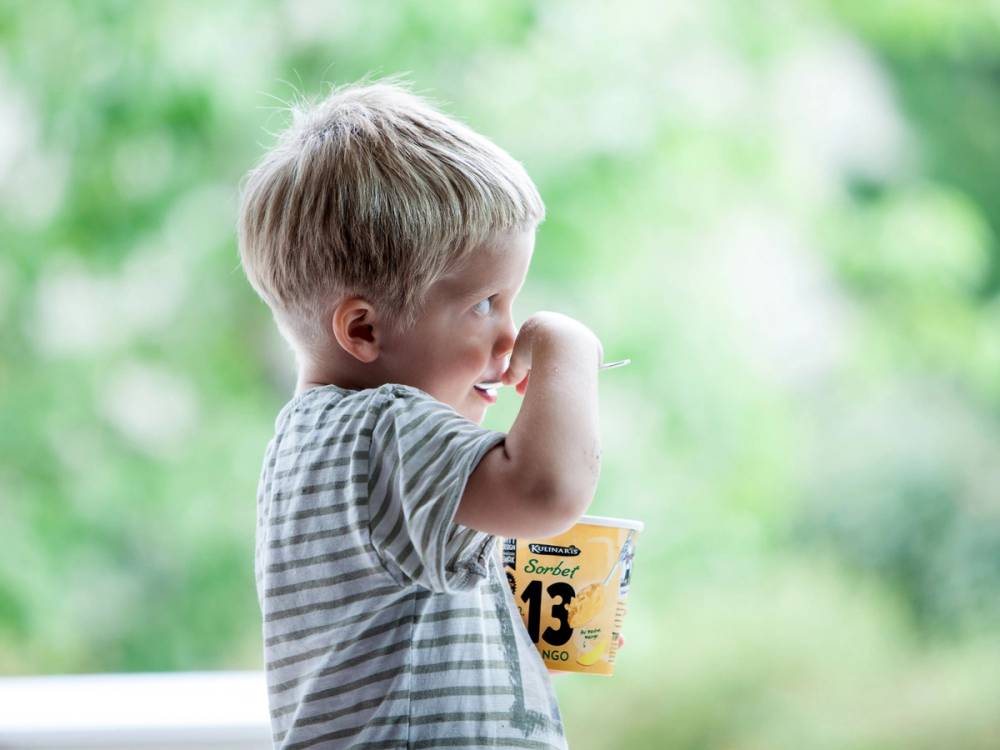 En ung gutt med kort blond hår som drikker av en kopp utendørs med grønt løv i bakgrunnen.