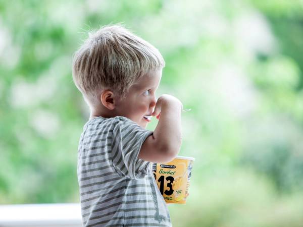 En ung gutt med kort blond hår som drikker av en kopp utendørs med grønt løv i bakgrunnen.