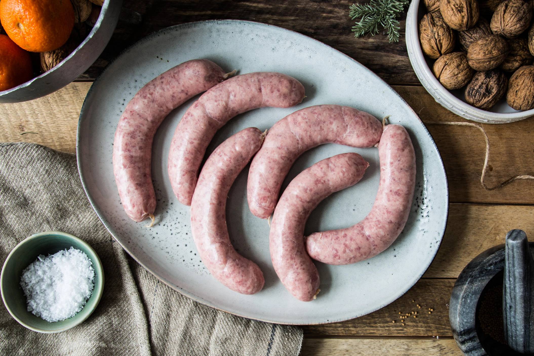 Fem julepolser av kjøtt ligger på et hvitt fat, omgitt av ulike krydder og nøtter på et trebord.