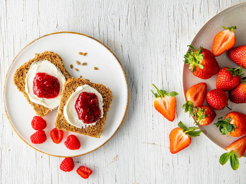 Bringebær og jordbær på en tallerken sammen med to brødskiver med jordbærsyltetøy og rømme, plassert på et hvitt bord.