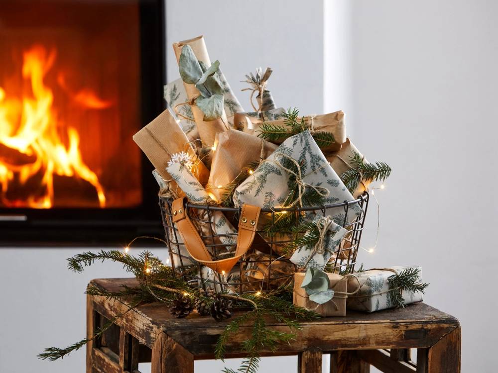 En kurv fylt med julegaver og dekorasjoner står på et bord foran en peis med tente flammer.