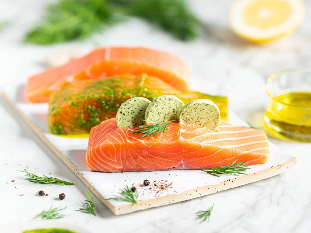 Lakseskiver med sitron, dill og grønne urter på en hvit skjærefjøl, med sitronbiter og olivenolje i bakgrunnen