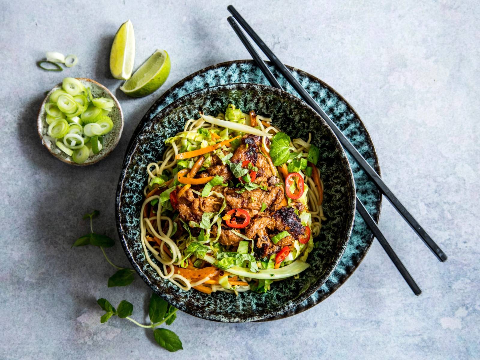En bolle med wokretter med grønnsaker og nudler, med spisepinner oppå, og sitronbåter ved siden av.