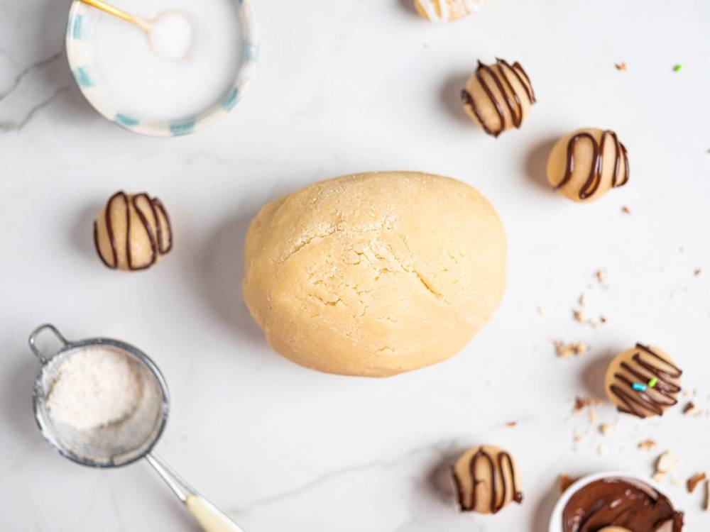 Marispandeig omgitt av små sjokoladetrukne kuler og en måleskje med mel på en hvit bakgrunn