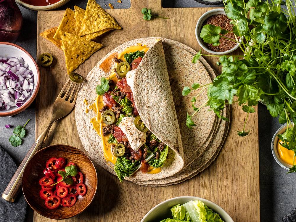 En burrito fylt med kjøttdeig, grønnsaker og ost på en tallerken, omgitt av små boller med tilbehør og grønne urter.