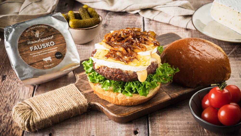 En burger med kjøtt, ost, salat og tomater på en treplate, med en pakke med Fausko brød i bakgrunnen.