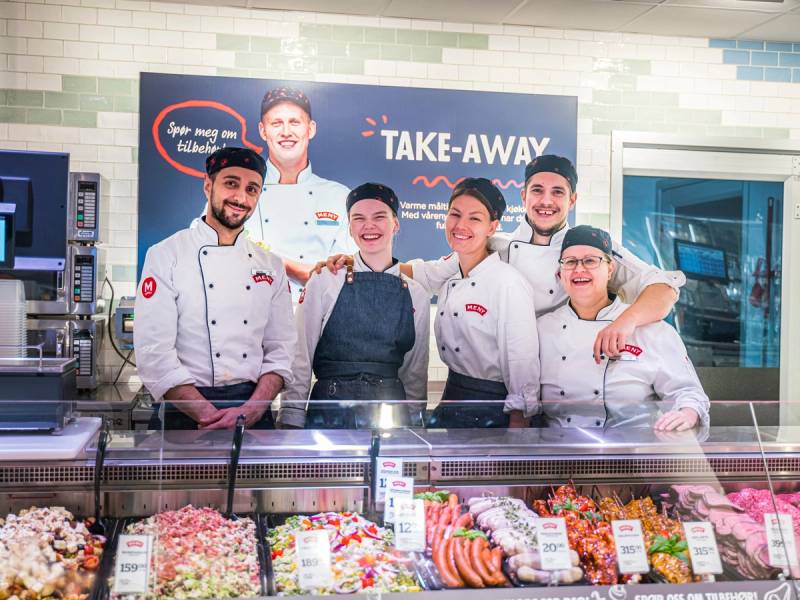 Gruppemedarbeidere i hvite kokkeklær bak disken på en matbutikk eller restaurant, med en kunde i midten, foran et skilt som sier 'TAKE-AWAY'.