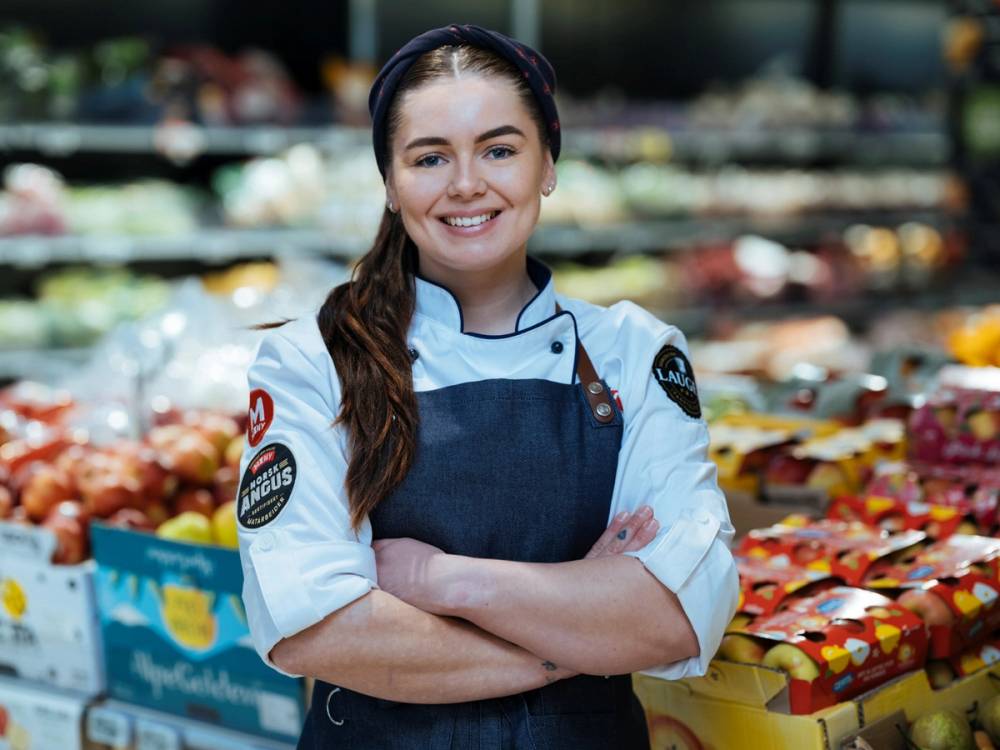 En person i kokkeforkle og uniform står med armene i kors foran en frukt- og grønnsaksavdeling i en butikk.