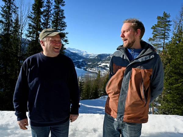 To personer står i snø med fjell og skog i bakgrunnen på en klar dag.