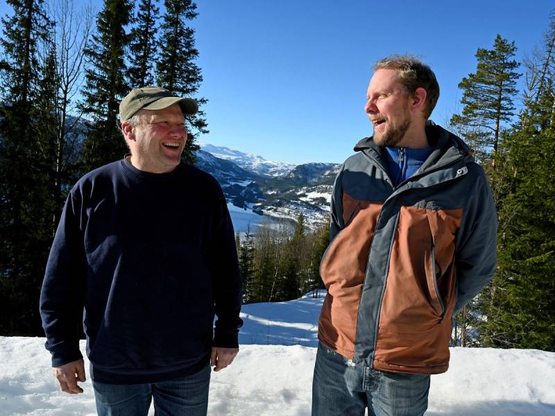 To personer står i snø med fjell og skog i bakgrunnen på en klar dag.