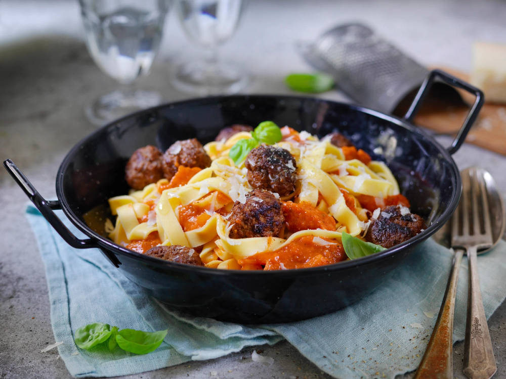 Pasta med kjøttboller i en svart skål, servert med grønne urter og tomatsaus.