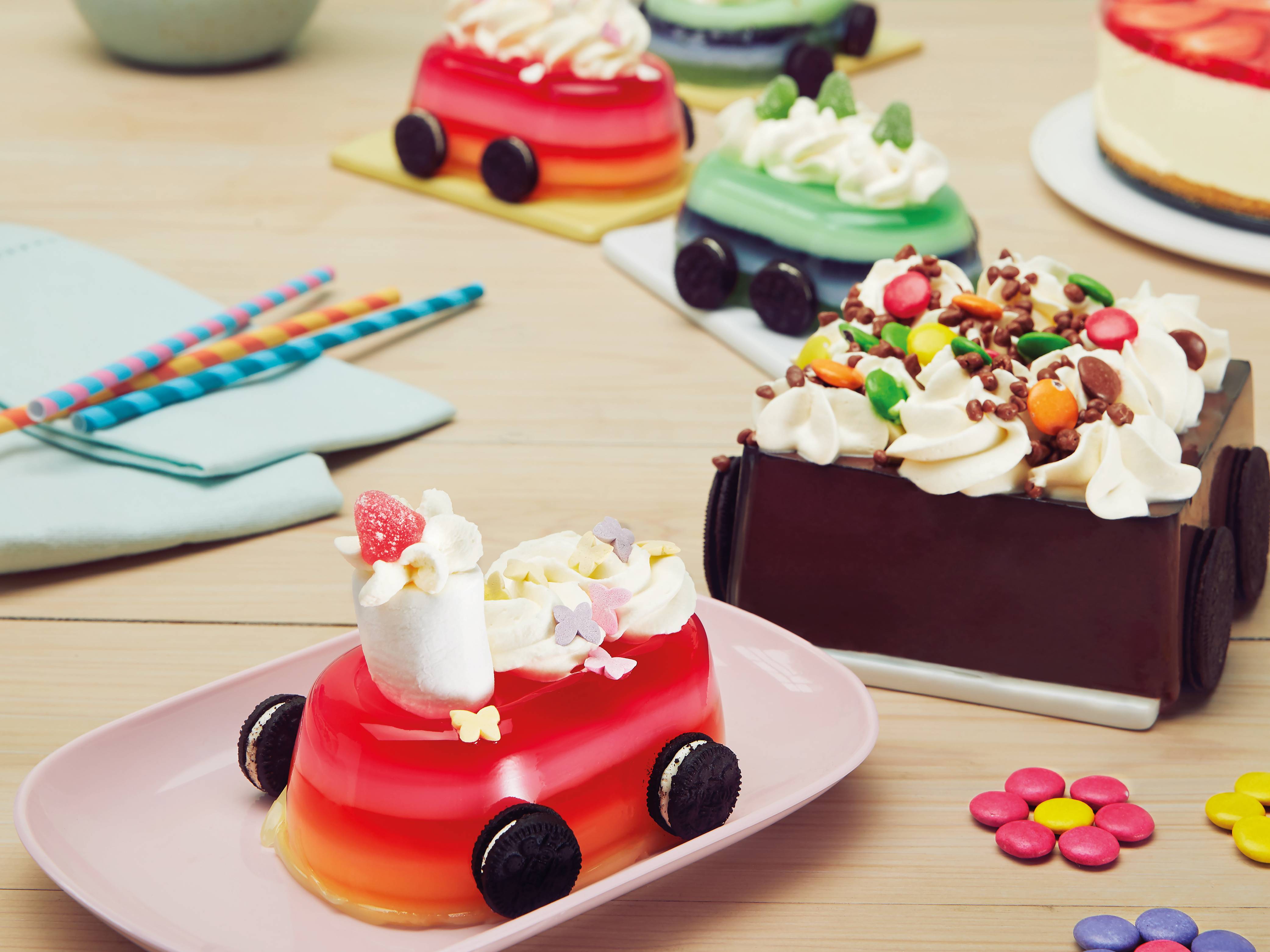 Fargerike kaker med ulike dekorasjoner og små pyntedetaljer på et bord, med noen lys og papirstrikker i bakgrunnen.