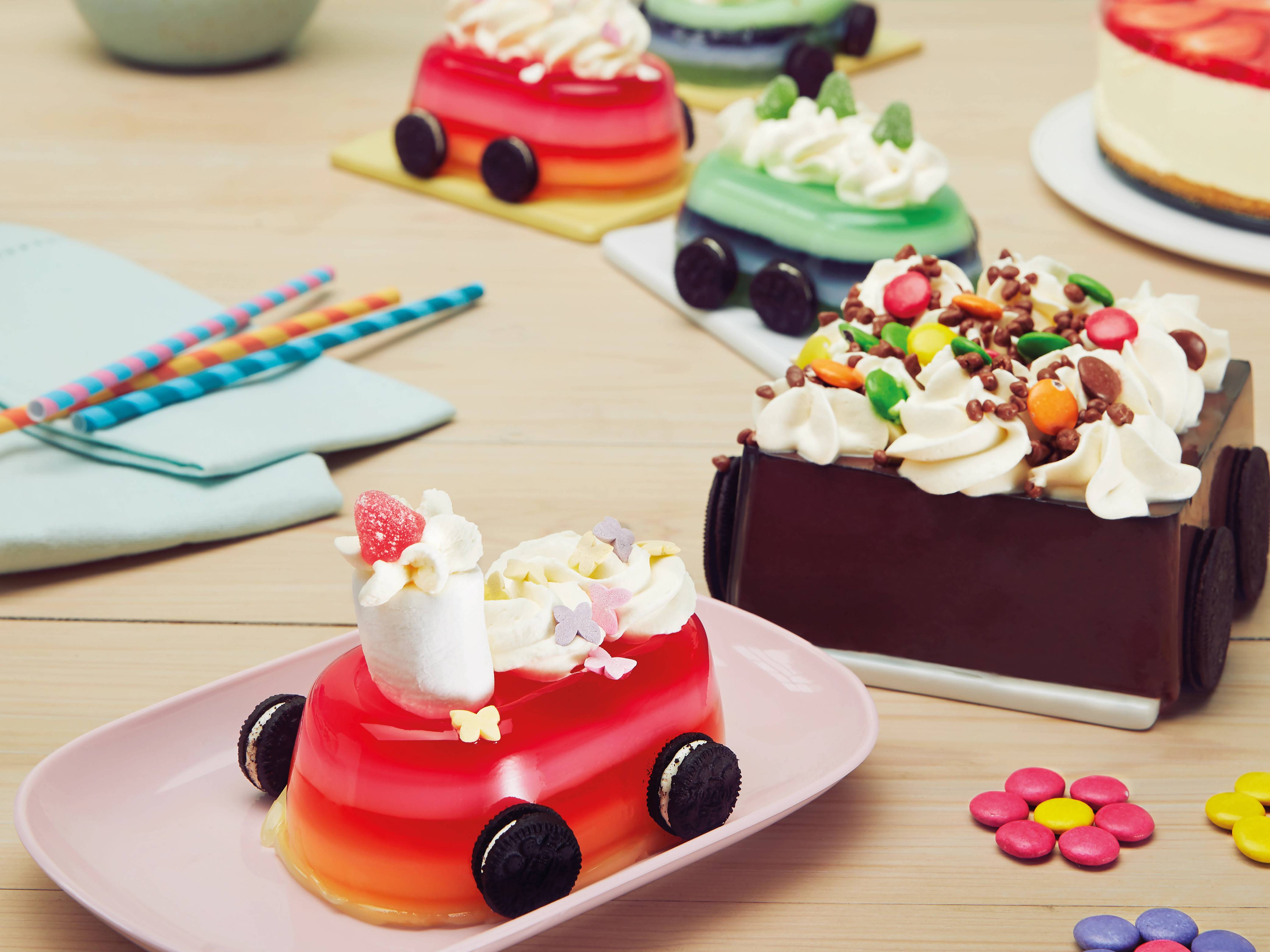 Fargerike kaker med ulike dekorasjoner og små pyntedetaljer på et bord, med noen lys og papirstrikker i bakgrunnen.