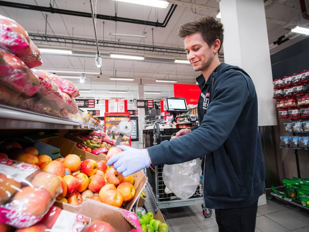 En mann handler frukt og grønt i en dagligvarebutikk, med fokus på fruktstativet foran ham.