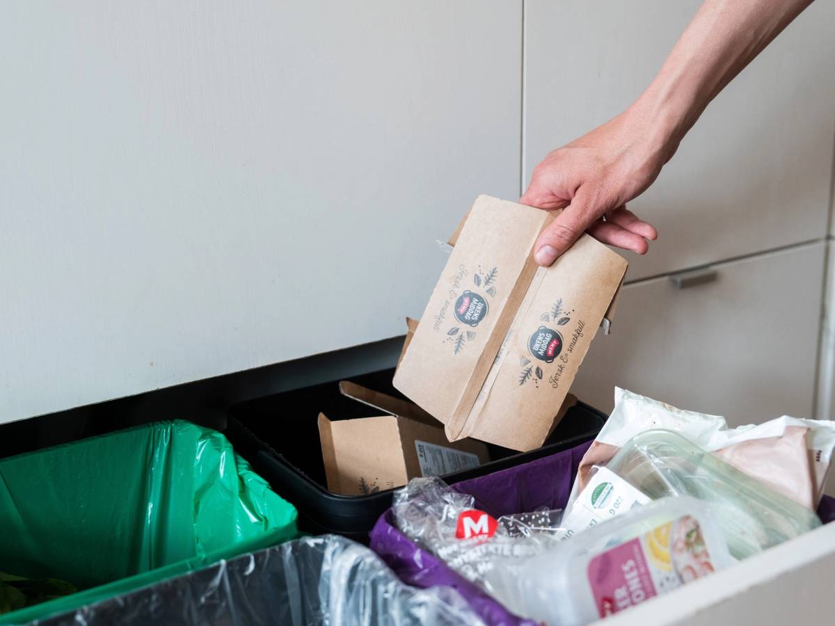 En person kaster matavfall i en søppelbøtte, med matemballasje og plastposer rundt.