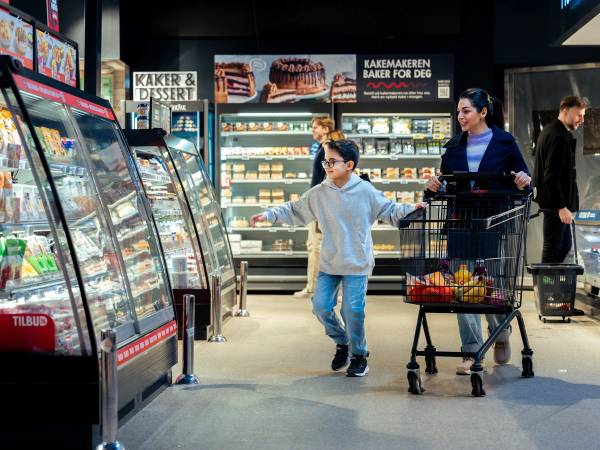 En familie med to barn handler i en dagligvarebutikk med hyller fylt av kaker og desserter i bakgrunnen.