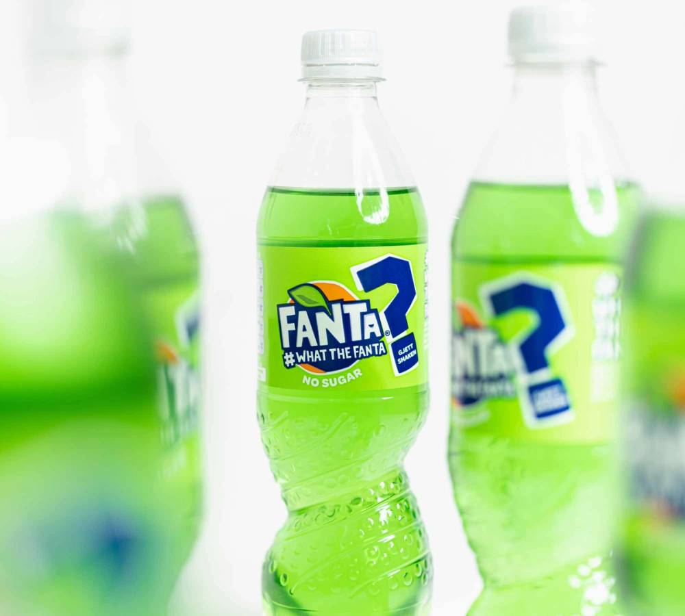 En flaske med Fanta-What the Fanta-etikett fylt med grønn drikke, omgitt av lignende flasker i bakgrunnen.