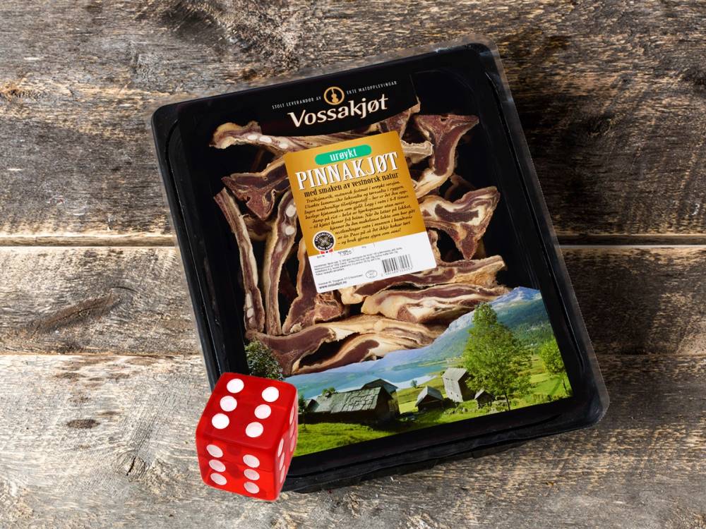 Pakke med Vossakjøtt med bilder av fjell og grønt landskap på emballasjen, plassert på en treflate med en rød terning foran.