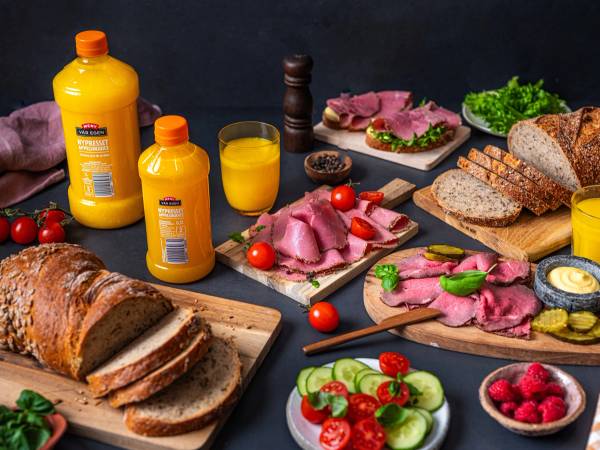 Et bord med ulike typer pålegg, brød, frukt og juice til lunsj, med en mørk bakgrunn.