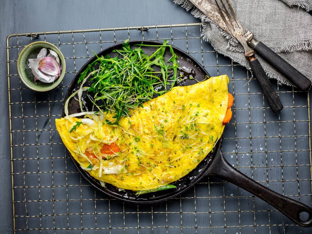 Omelett med ris og grønnsaker på en svart stekepanne, ser ut til å være servert med en liten skål med løk og urter ved siden av.