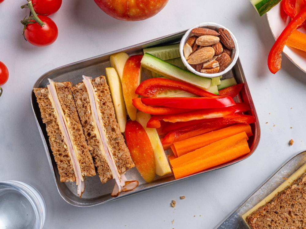 En sunn matpakke med små sandwiches, oppkuttede grønnsaker og en liten kopp med nøtter, på en svart tallerken.