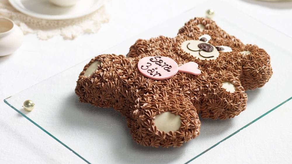 En sjokoladekake formet som en bjørn med pyntede øyne og munn, dekorert med sjokoladestrøssel og krem, plassert på et hvitt fat.