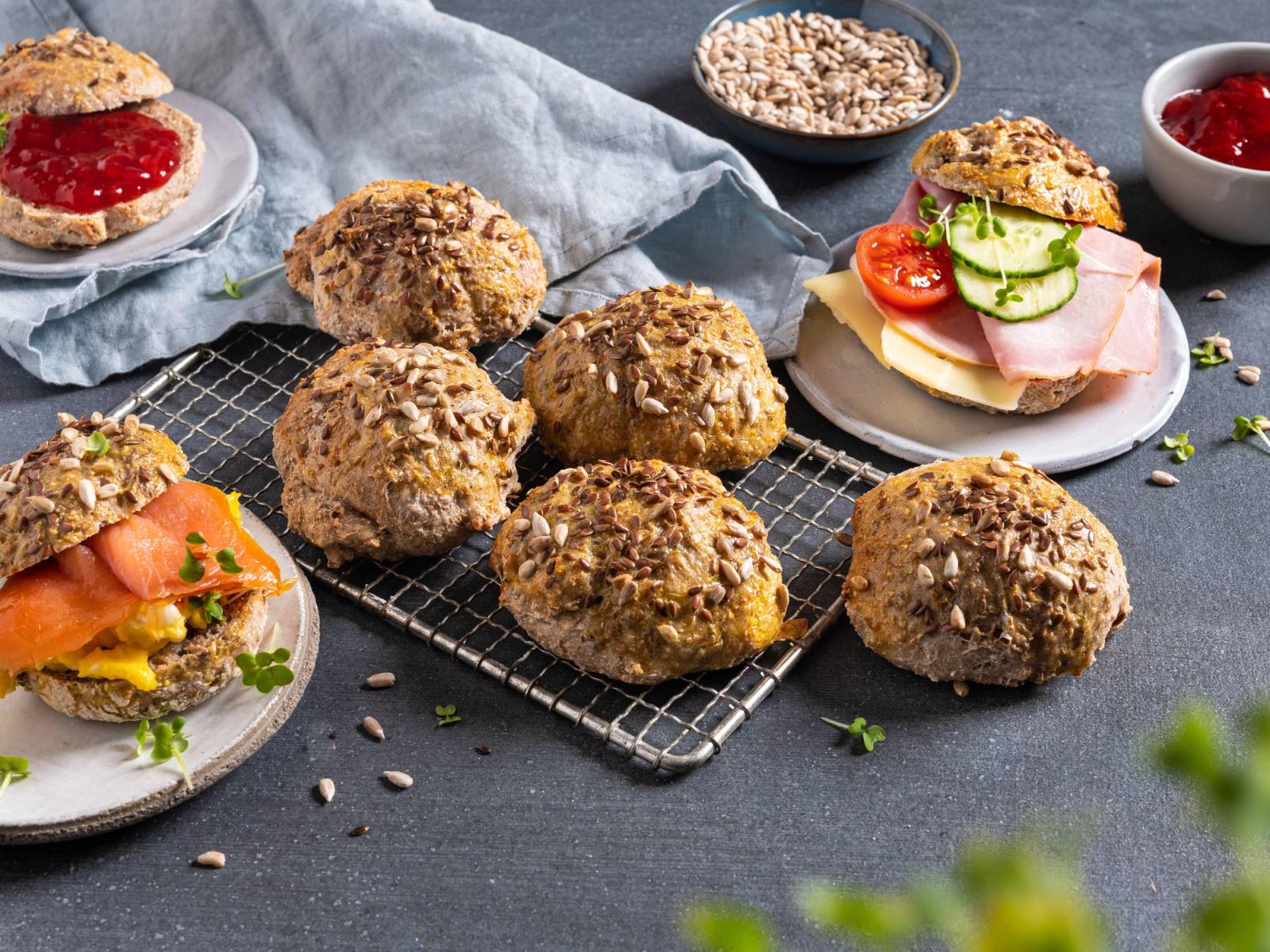 Grove, runde rundstykker på en avkjølingsrist med noen pålegg og tilbehør rundt, som syltetøy og skiver av grønnsaker og kjøttpålegg.