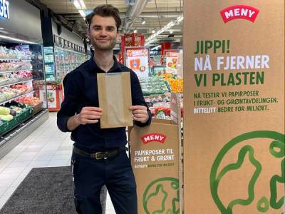 Person som står i en butikk med en pappeske, omringet av plastemballasje og reklameskilt for plastgjenvinning.
