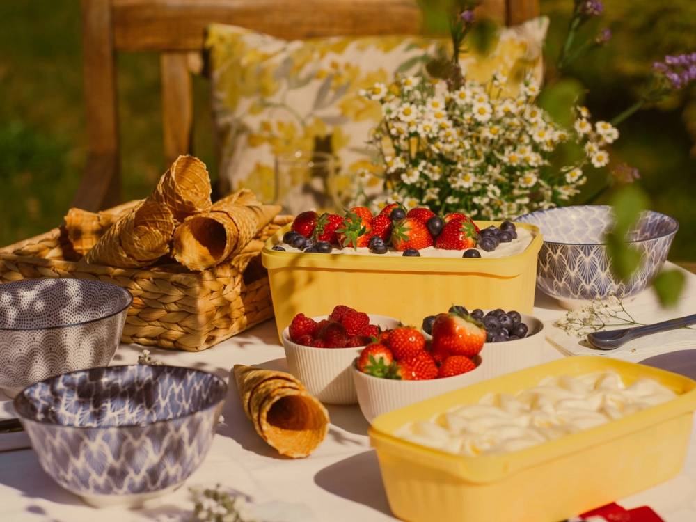 Frukt og bær på et bord med blomster og kake, sannsynligvis en utendørs piknik eller fest, i en hage eller park.