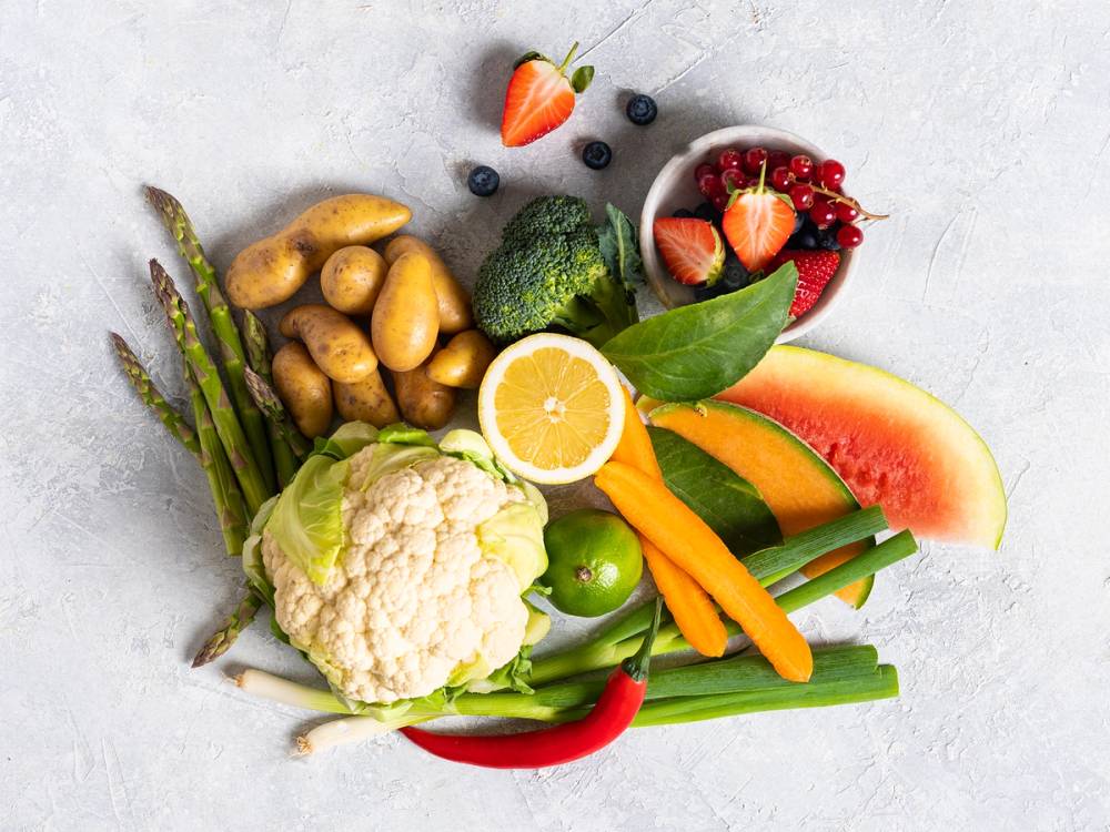 Frukt og grønnsaker som oppbevares i en åpen bolle og på tallerken, inkludert jordbær, blåbær, sitron, vannmelon, agurk, paprika, brokkoli, asparges, og poteter.