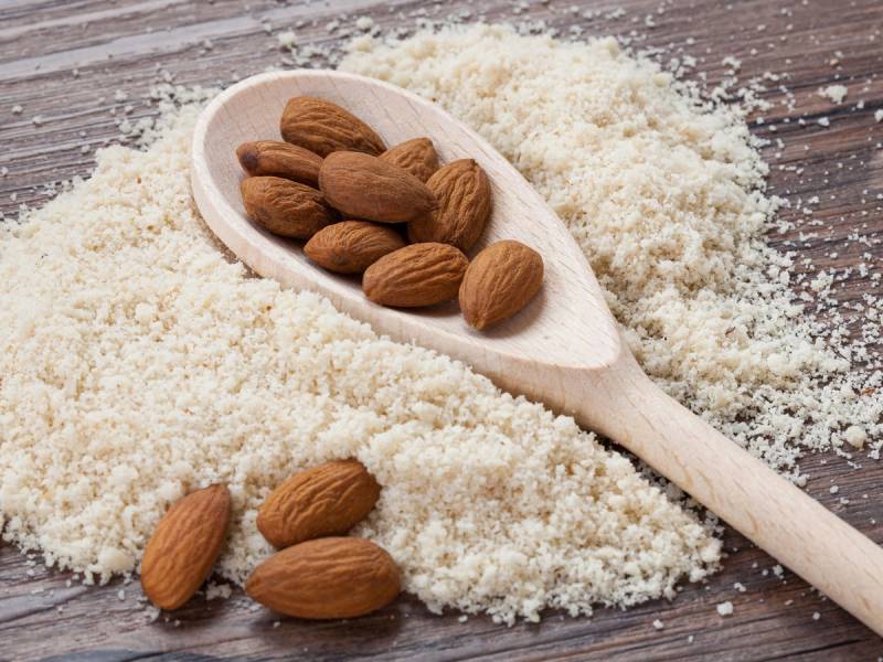 Håndfull med mandler på en skje, omgitt av glutenfritt mel på et treverk, brukt til baking.