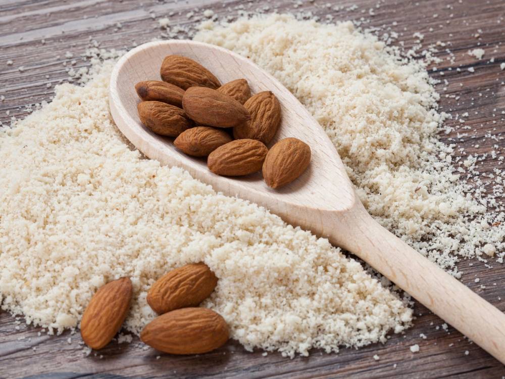 Håndfull med mandler på en skje, omgitt av glutenfritt mel på et treverk, brukt til baking.