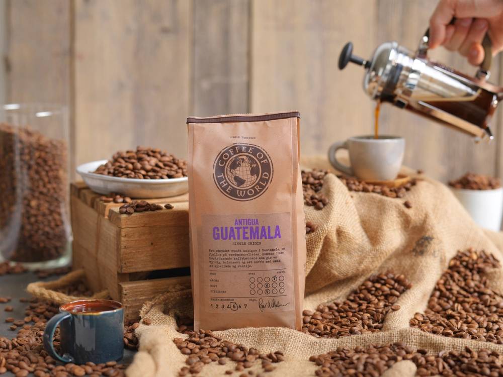 Kaffe fra Antigua Guatemala er presentert med en pose av månedens kaffe, omgitt av kaffebønner og en kopp, med en barista som heller kaffe i bakgrunnen.