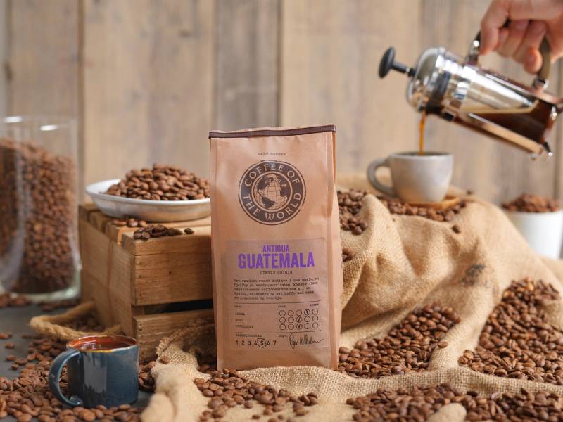 Kaffe fra Antigua Guatemala er presentert med en pose av månedens kaffe, omgitt av kaffebønner og en kopp, med en barista som heller kaffe i bakgrunnen.