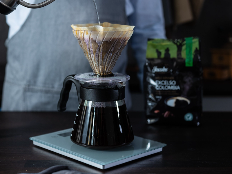 Håndbrygging av kaffe i en glasskanne på en elektrisk kaffebrygger, med en espressobønnepakke i bakgrunnen.