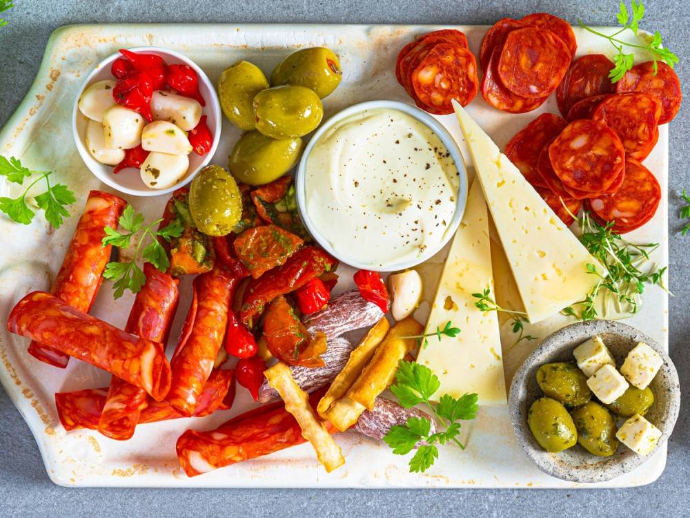 Et fat med ulike typer tapas, inkludert oliven, cherrytomater, ost, og spekepølser, pyntet med frisk urter.