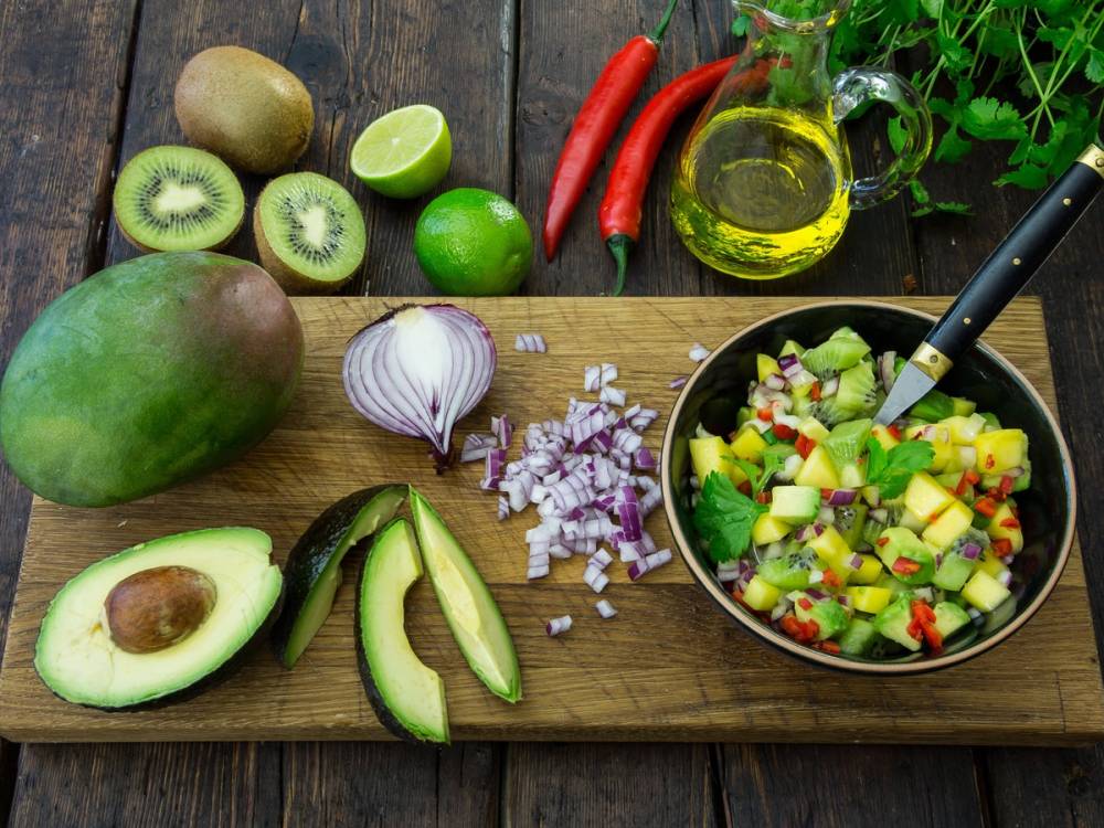 Skjærebrett med avokado, løk, lime, kiwi, chili, olje og en salat med agurk og frukt, samt en bolle med salat og en kniv, på et treverk.